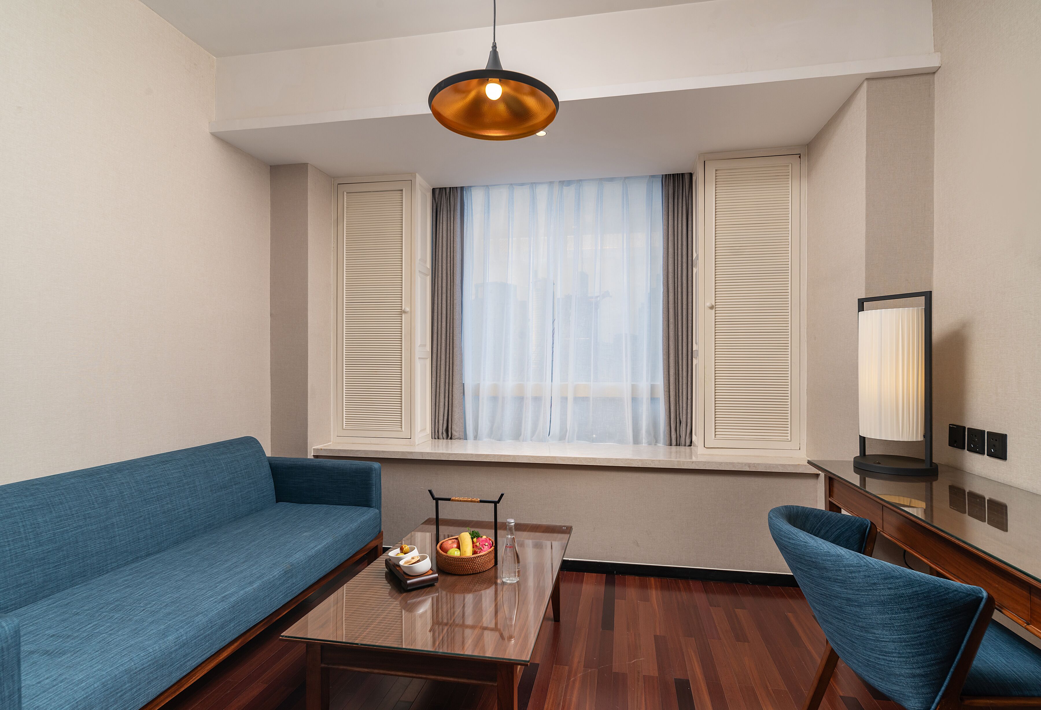 A modern hotel room with a blue sofa, a wooden coffee table with fruit, a hanging light fixture, and a large window with blinds.