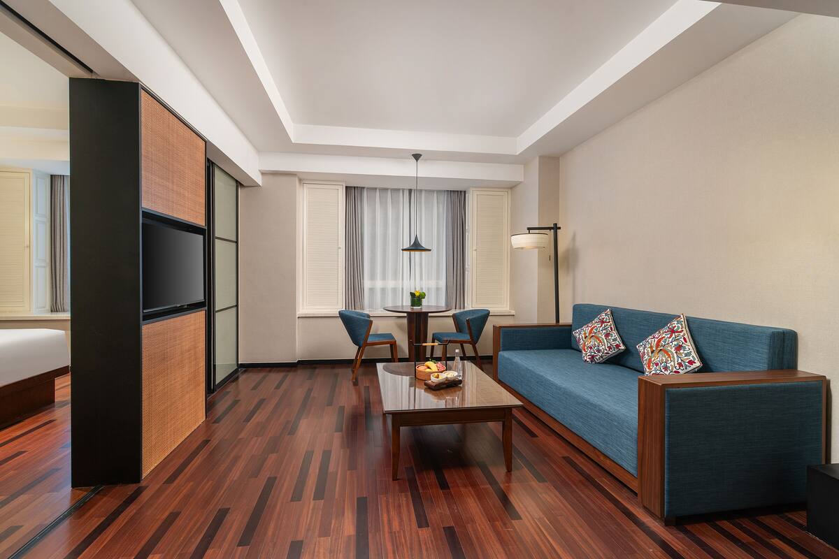 Een moderne hotelkamer met een houten vloer, een blauwe bank met kleurrijke kussens, een klein tafeltje met fruit en een televisie op een zwarte standaard.