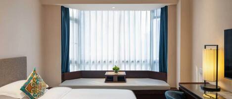 A hotel room with a bed, a desk, a chair, a lamp, and a window with curtains.