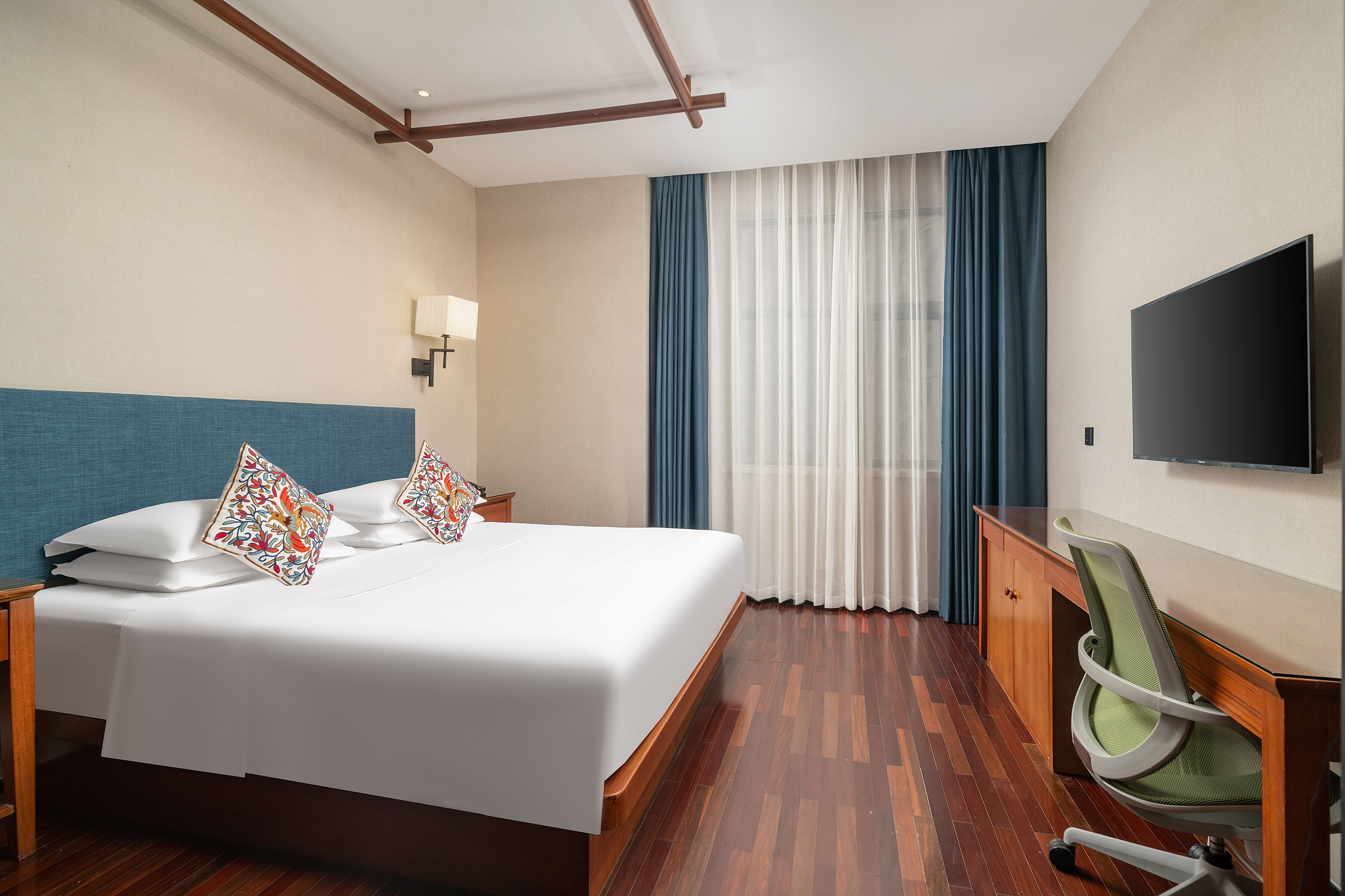 Quarto de hotel com cama, escrivaninha, cadeira, televisão e janela com cortinas.