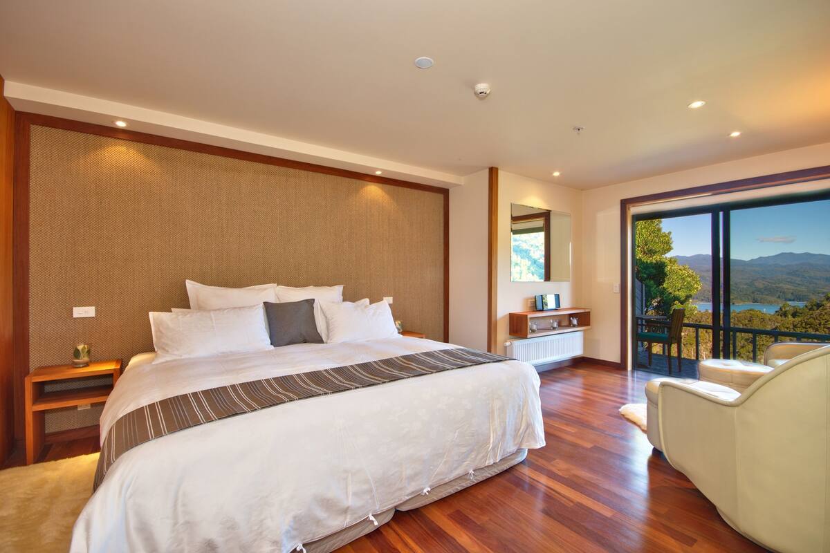 A modern bedroom with a large bed, a nightstand, a chair, and a view of the outdoors.