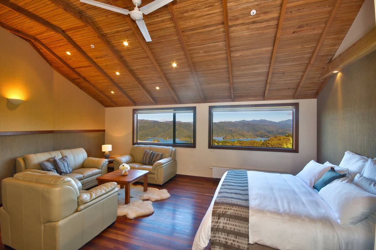 A spacious room with a wooden ceiling, a large bed, two sofas, and a view of mountains through the windows.