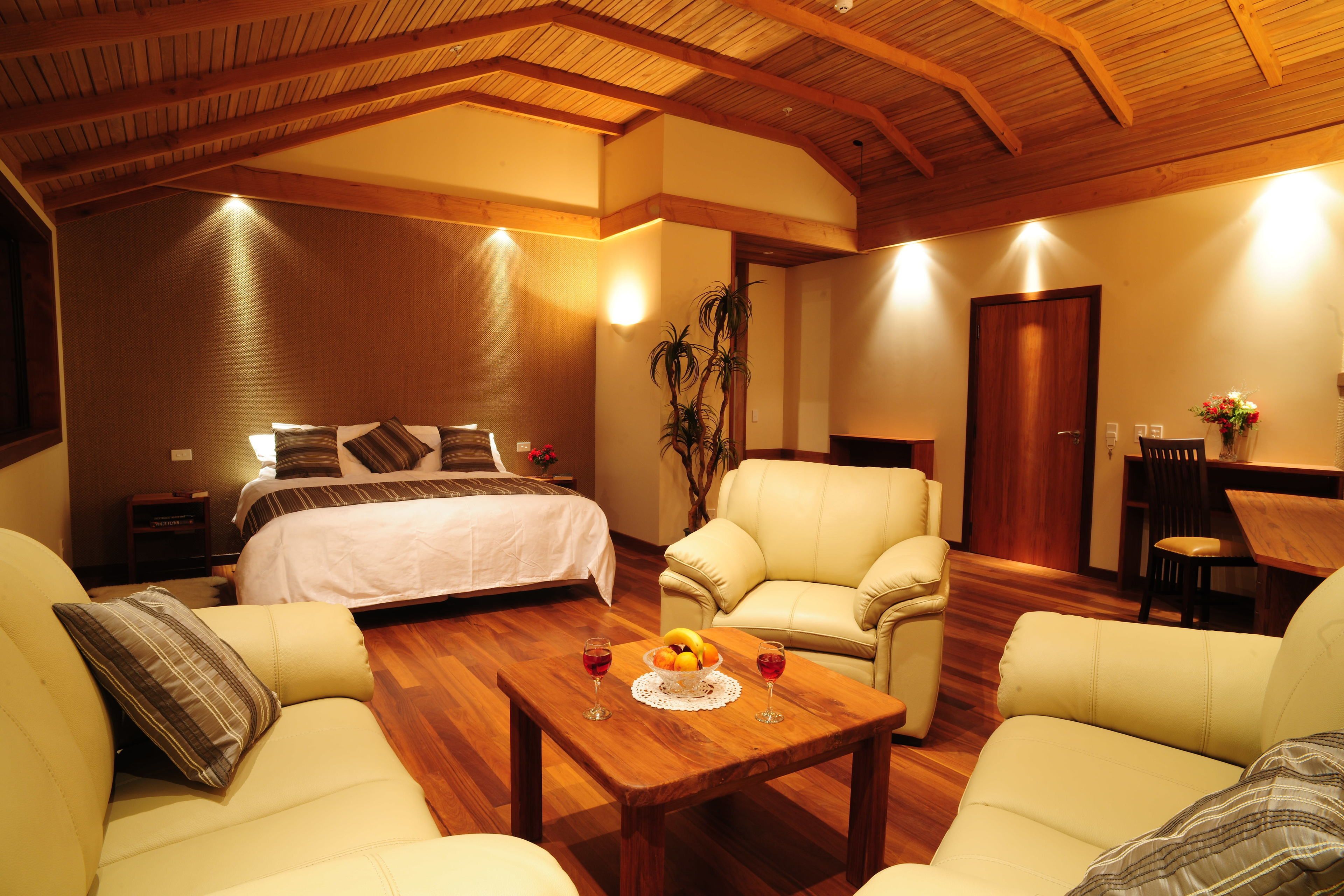 A hotel room with a bed, two sofas, a wooden coffee table with fruit and wine glasses, a dining table, and a plant.