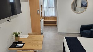 Habitación de hotel con cama, escritorio con silla, una mesita con una planta, microondas, refrigerador, televisor y ventana con cortinas.