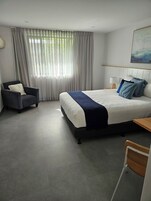 Una habitación de hotel moderna con una cama grande, una silla, una mesita de noche y un ventanal con cortinas.