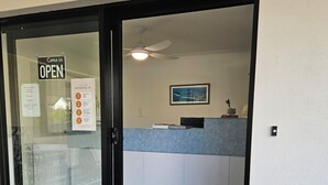 Zona de recepción de un hotel con puerta de cristal, letrero que indica que el establecimiento está abierto y mostrador de recepción con una computadora y un cuadro en la pared.