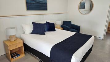 A modern hotel room with a large bed, a bedside table with a lamp, a chair, a round mirror, and a painting on the wall.