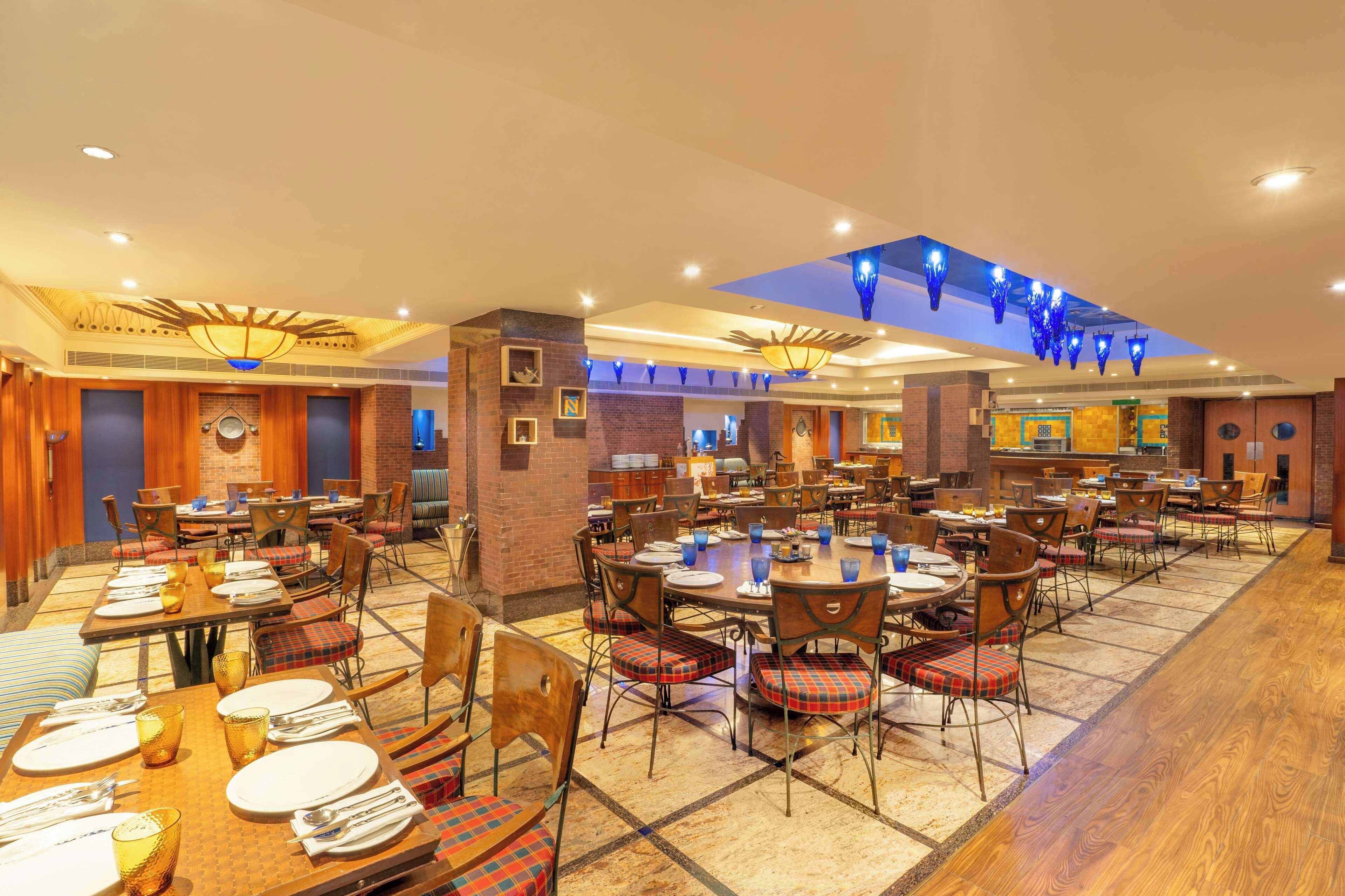 A spacious dining area with multiple tables and chairs, a brick wall, and a ceiling with blue decorative lights.
