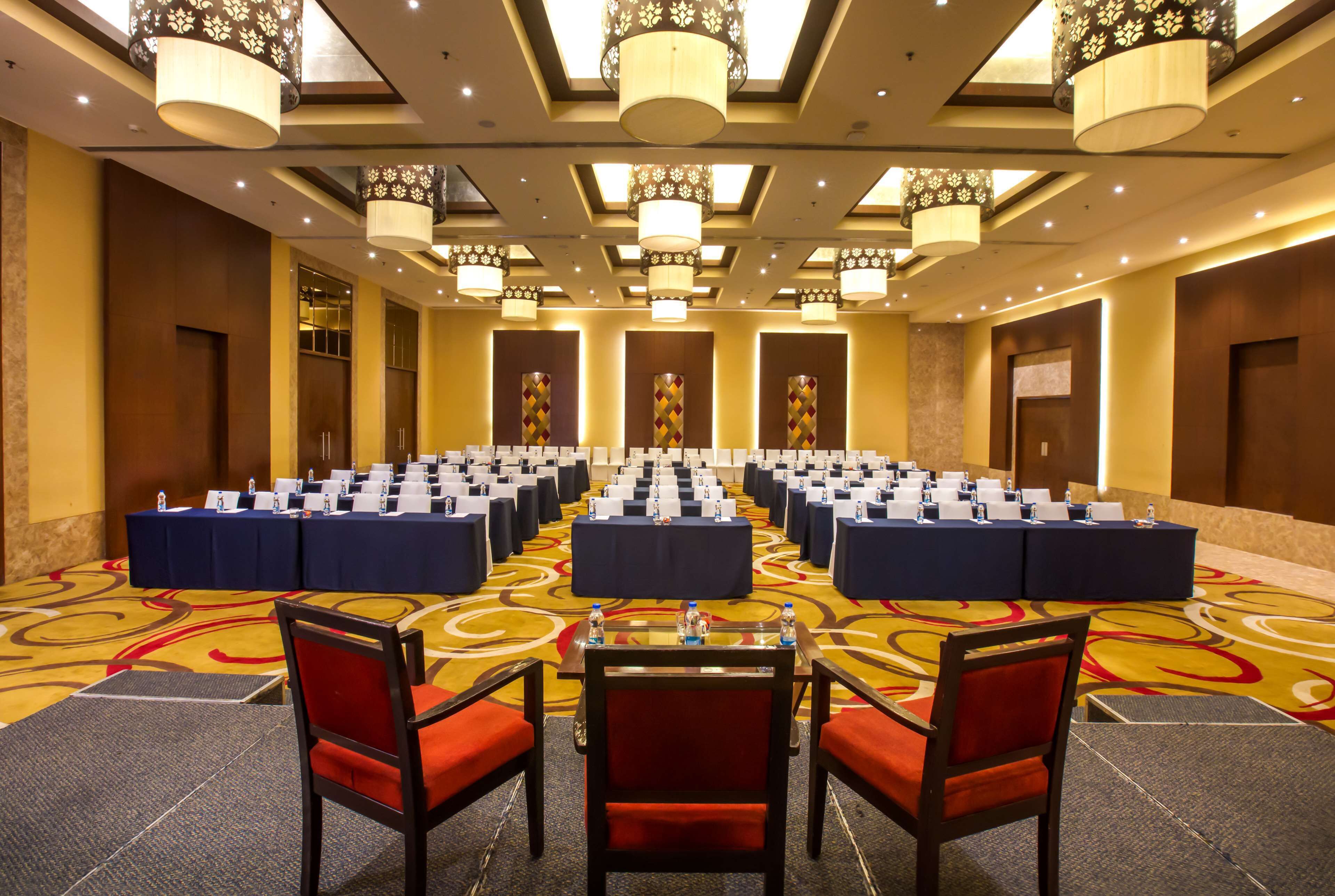 A conference room with rows of tables and chairs, large chandeliers, and a patterned carpet.