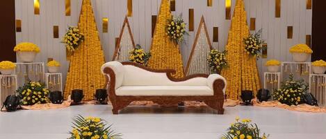 Un escenario con un sofá blanco, arreglos florales amarillos y un fondo blanco con líneas verticales.