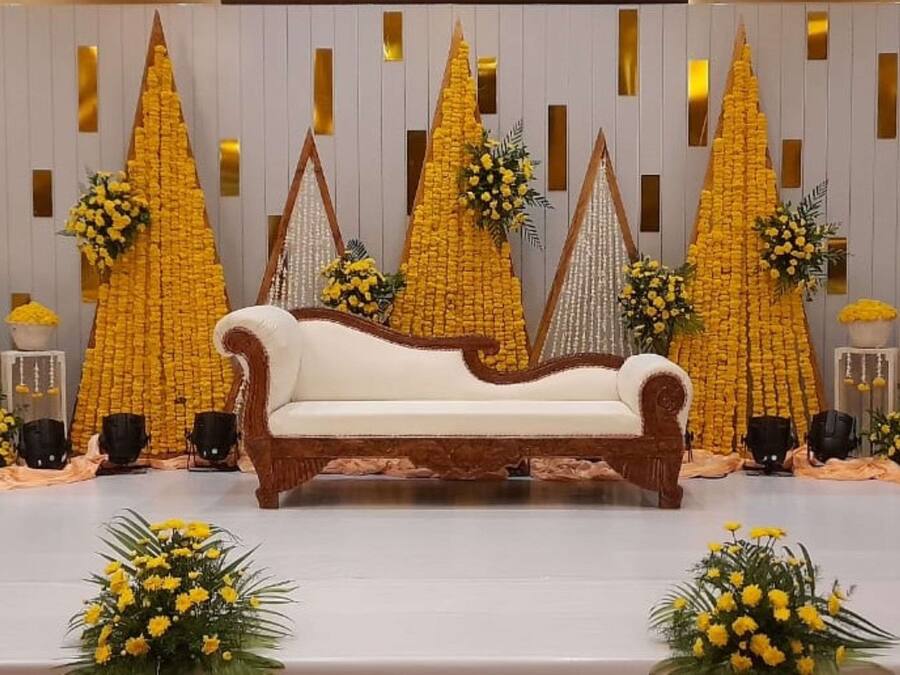 Un escenario con un sofá blanco, arreglos florales amarillos y un fondo blanco con líneas verticales.