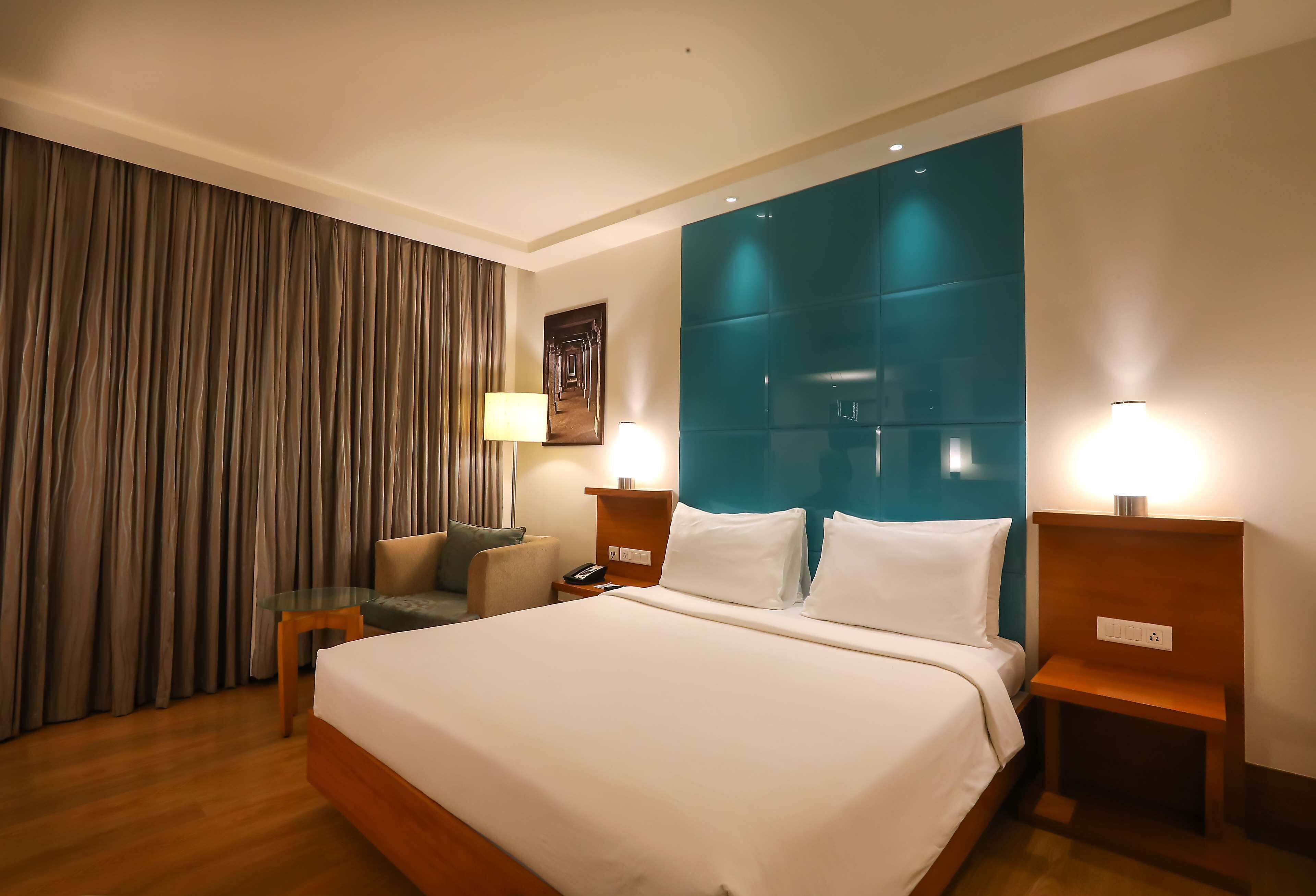 A hotel room with a large bed, a nightstand with a lamp, a seating area with a chair and a sofa, and a curtain on the window.