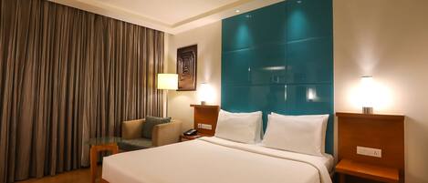 A hotel room with a large bed, a nightstand with a lamp, a seating area with a chair and a sofa, and a curtain on the window.