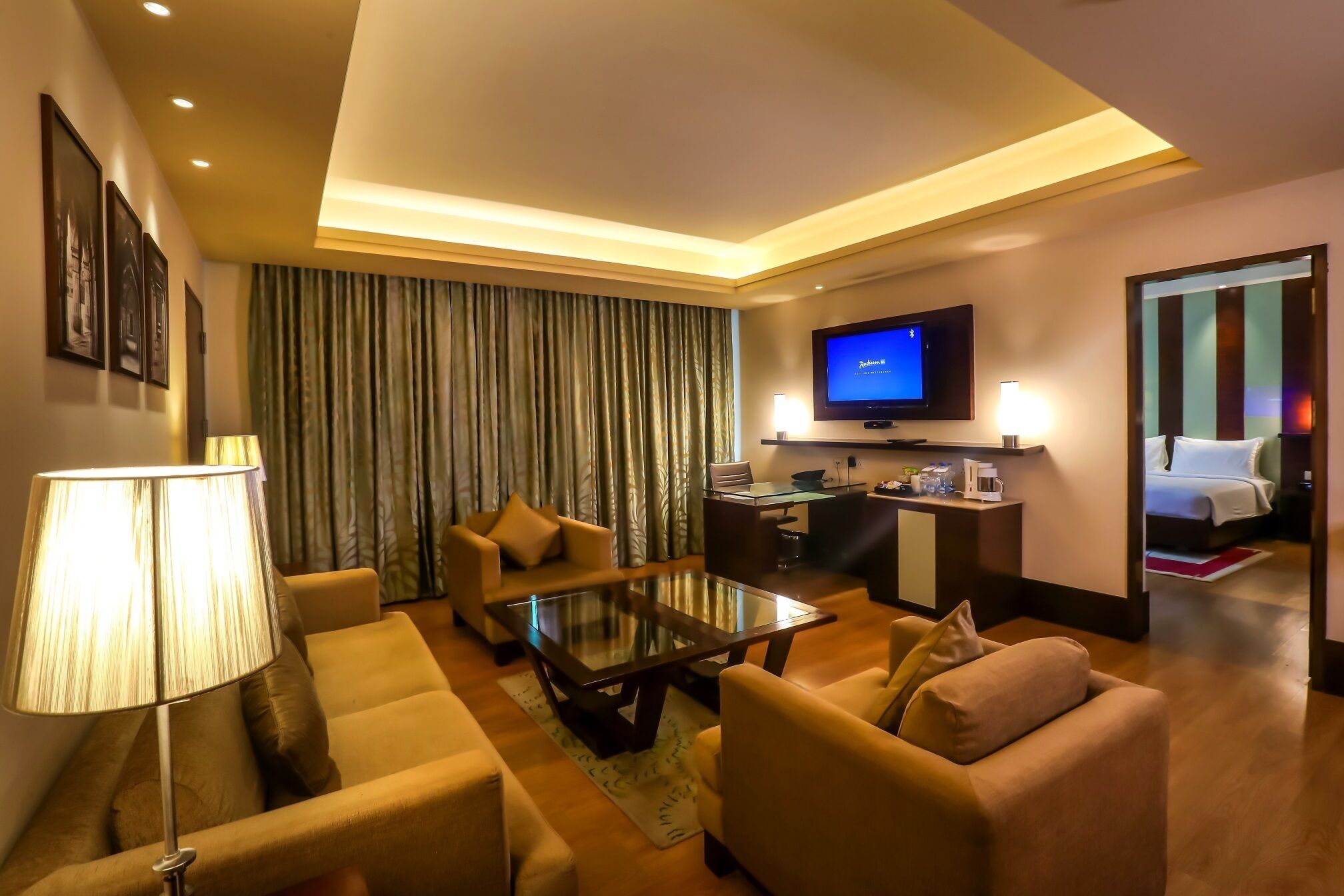 Habitación de hotel con sala de estar que incluye un sofá, una mesa de centro de vidrio, un televisor de pantalla plana y una lámpara.