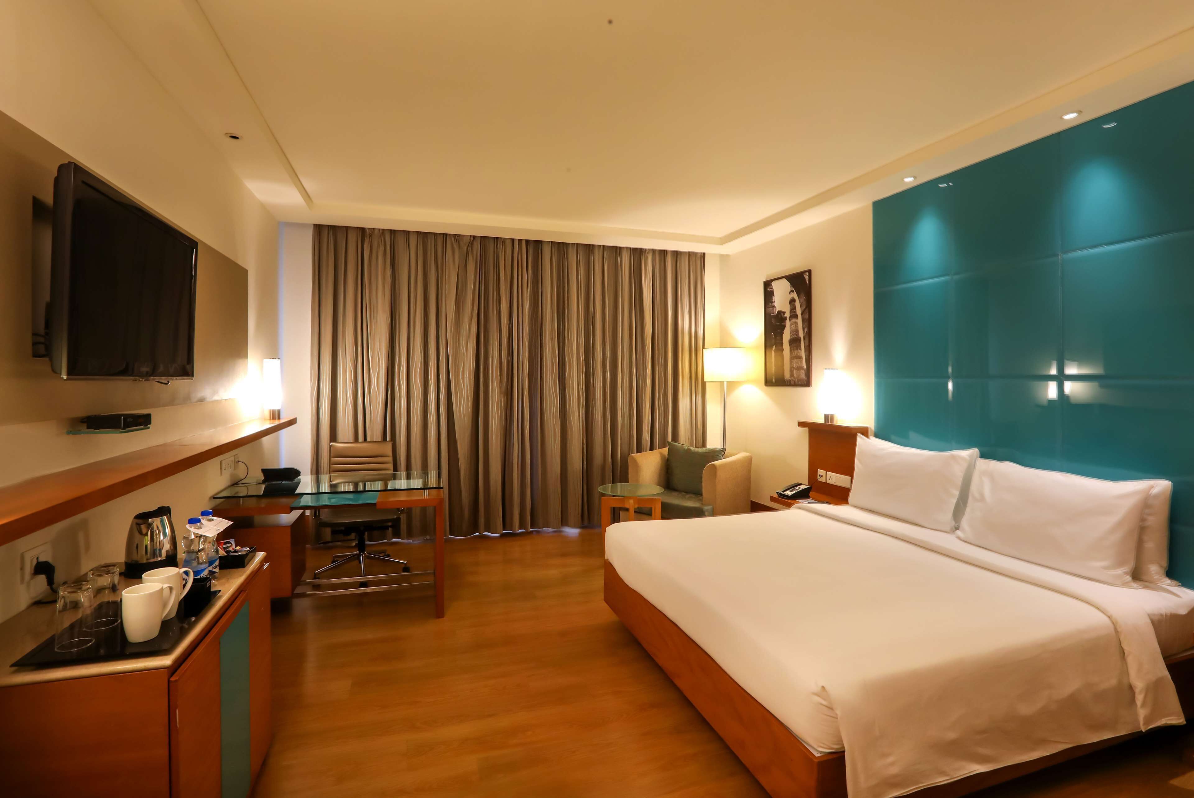 Habitación de hotel con cama, escritorio con silla, televisor montado en la pared, ventana con cortinas y una pared de acento azul.