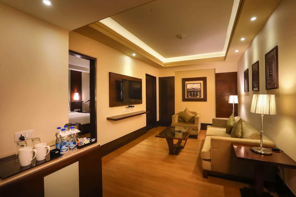 Ein Hotelzimmer mit einer Couch, einem Couchtisch, einem an der Wand montierten Fernseher, einer Lampe und einem kleinen Tisch mit Wasserflaschen und Bechern.