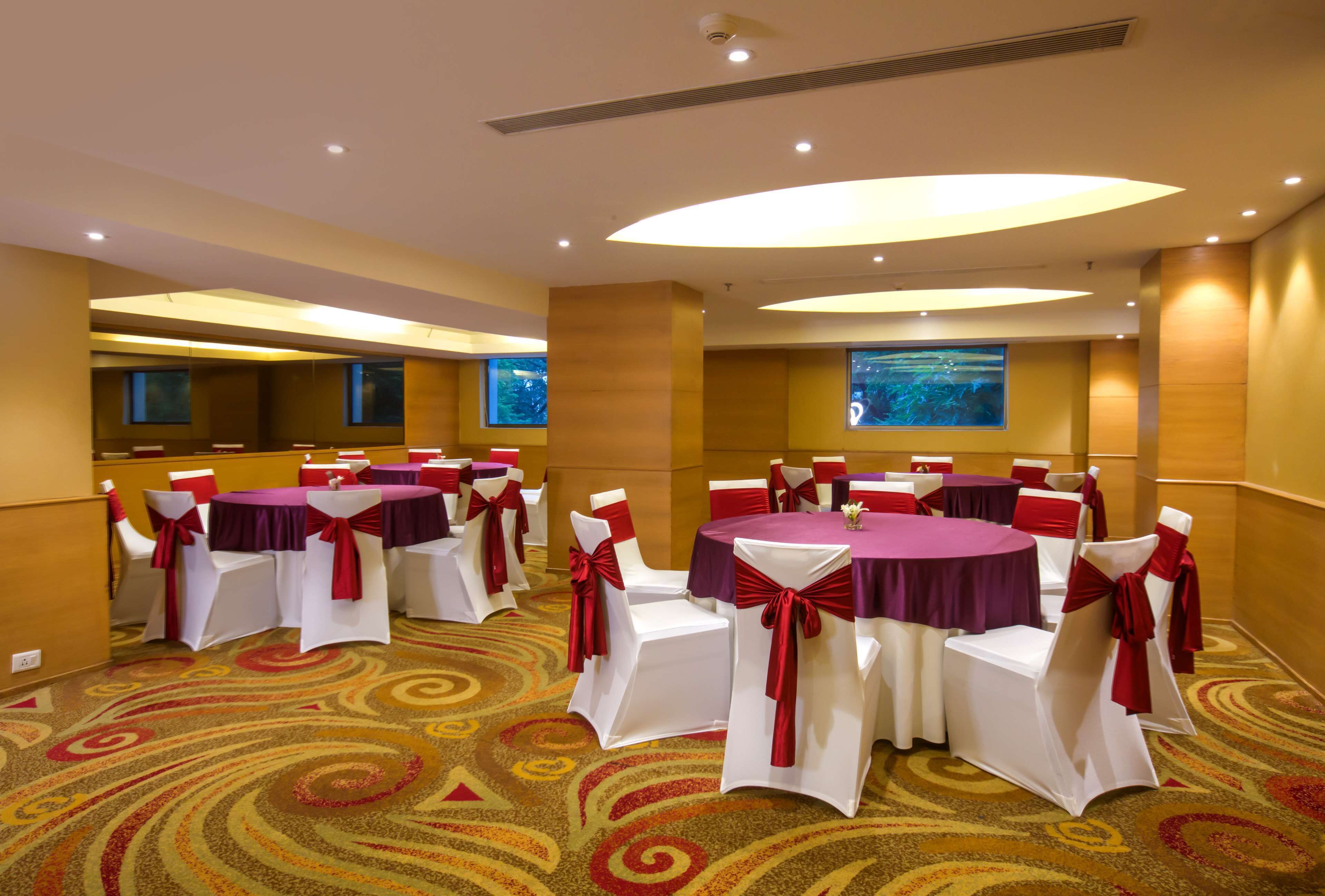 A conference room with round tables and white chairs, each with red sashes. The room has a carpeted floor and a large window with a view.