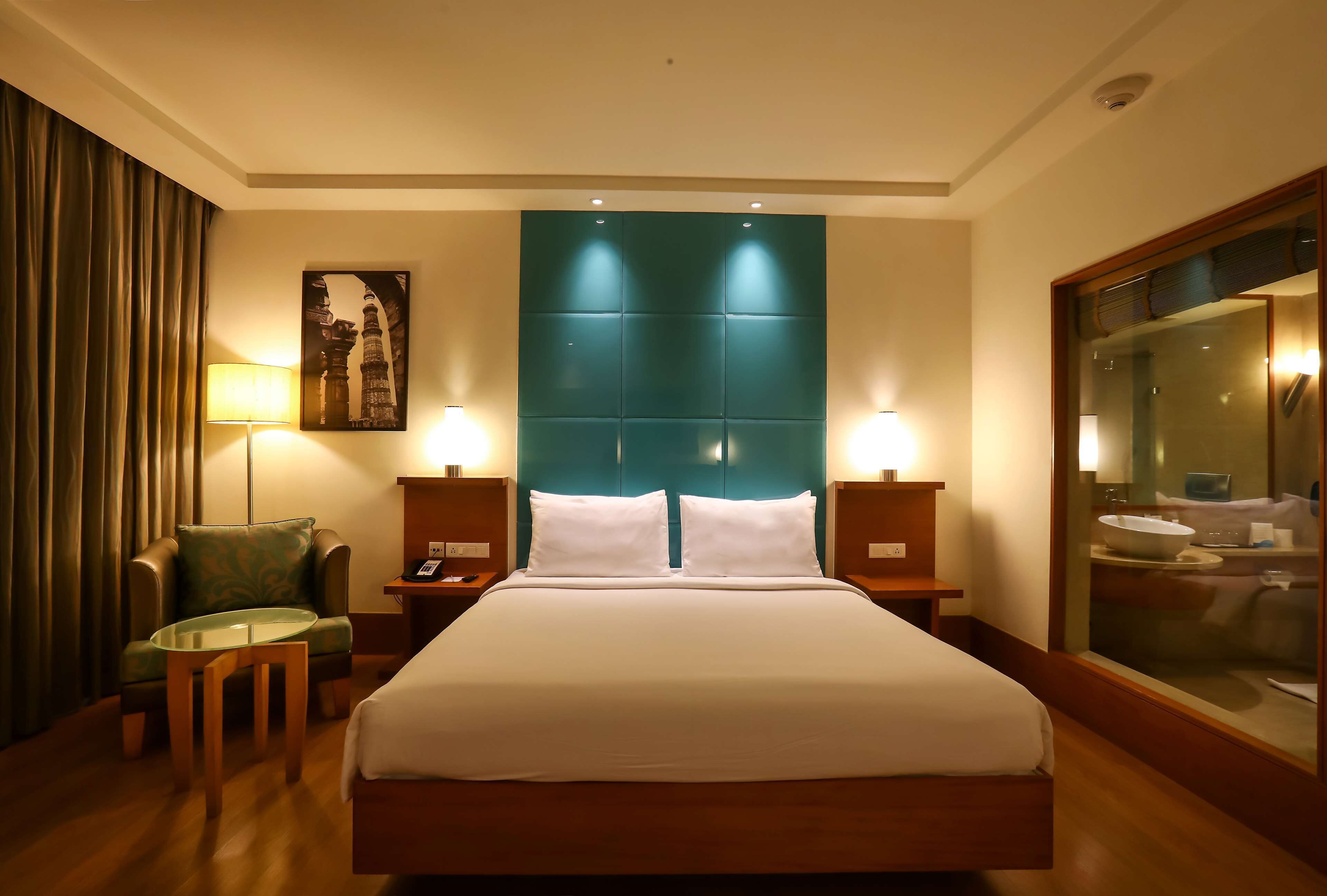 A hotel room with a large bed, two bedside tables with lamps, a green armchair, a small table, and a large mirror.