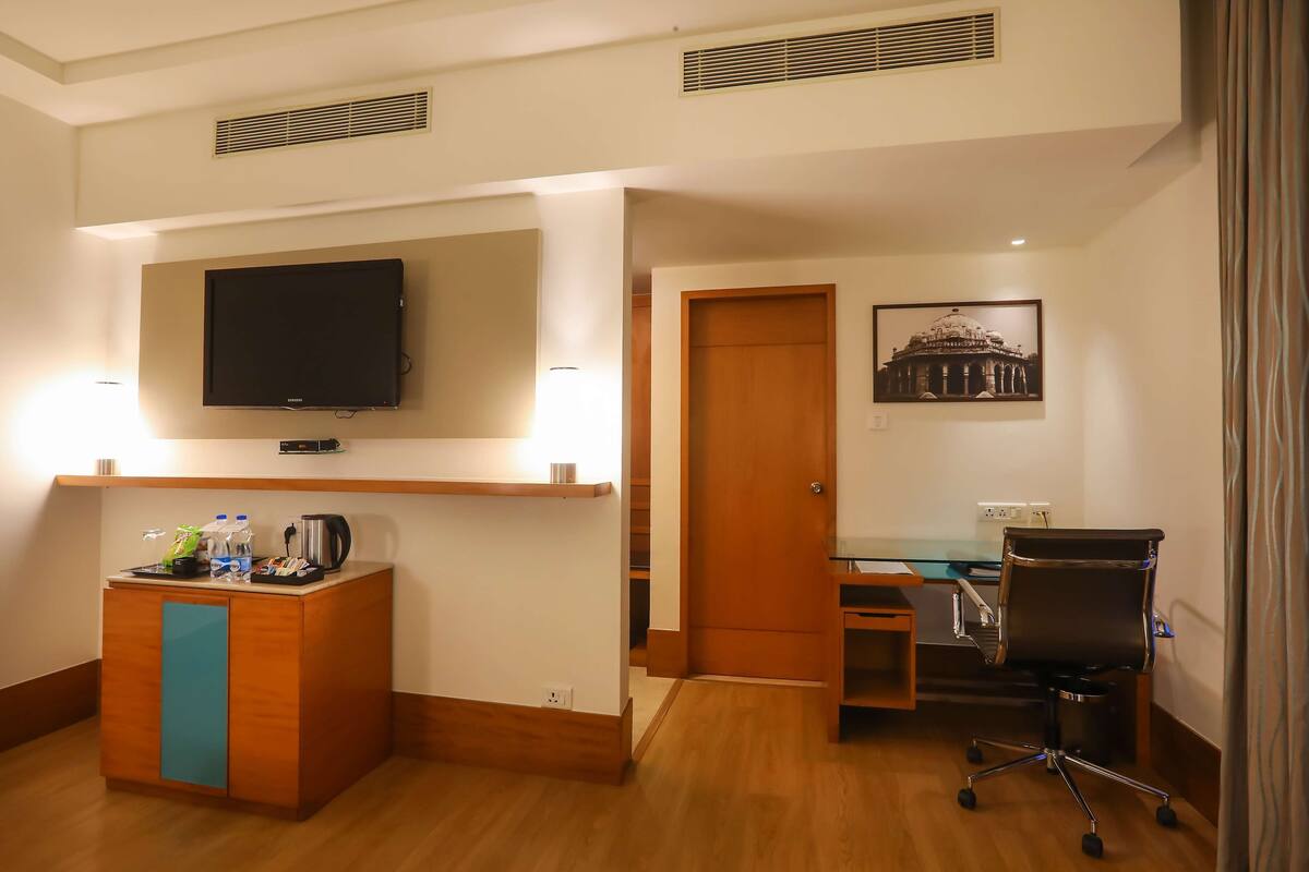Ein Hotelzimmer mit Schreibtisch aus Holz, schwarzem Bürostuhl, Wandfernseher, kleinem Empfangstresen mit blauer Verkleidung und einem gerahmten Bild an der Wand.