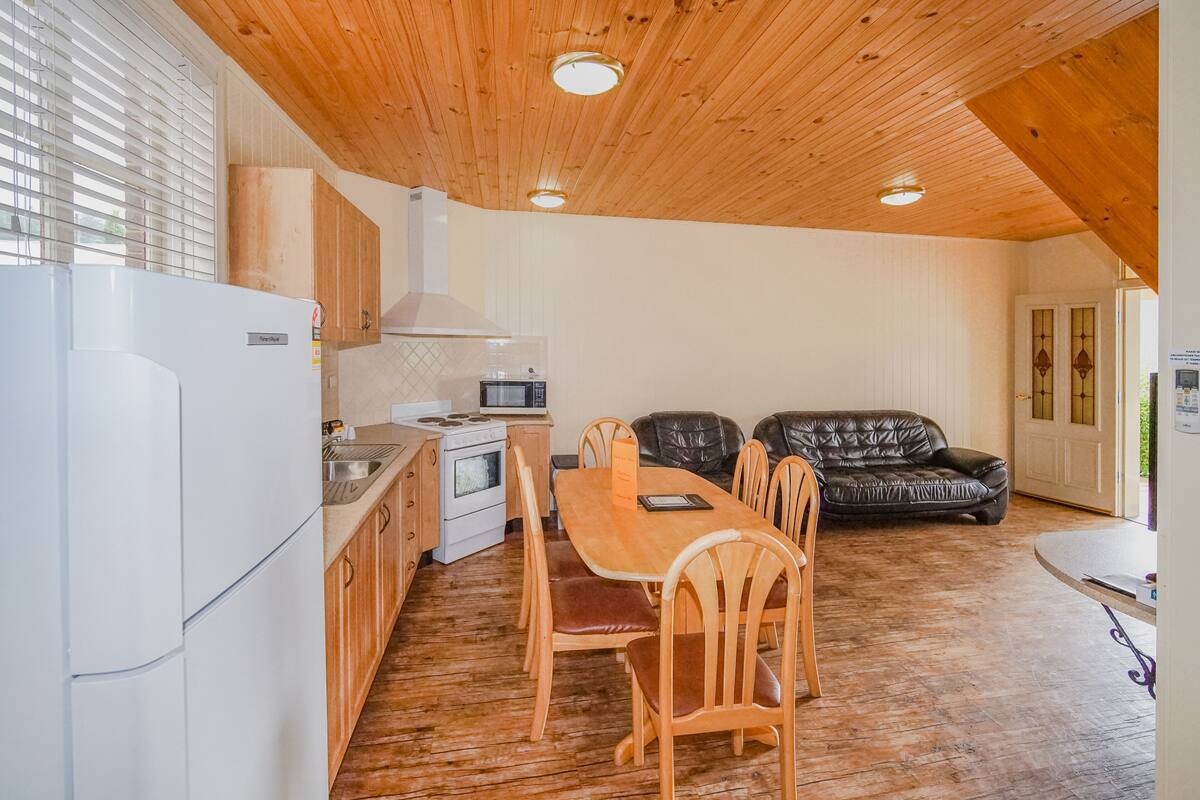 A compact kitchen with wooden cabinets, a dining table with chairs, a black leather sofa, and a wooden ceiling.