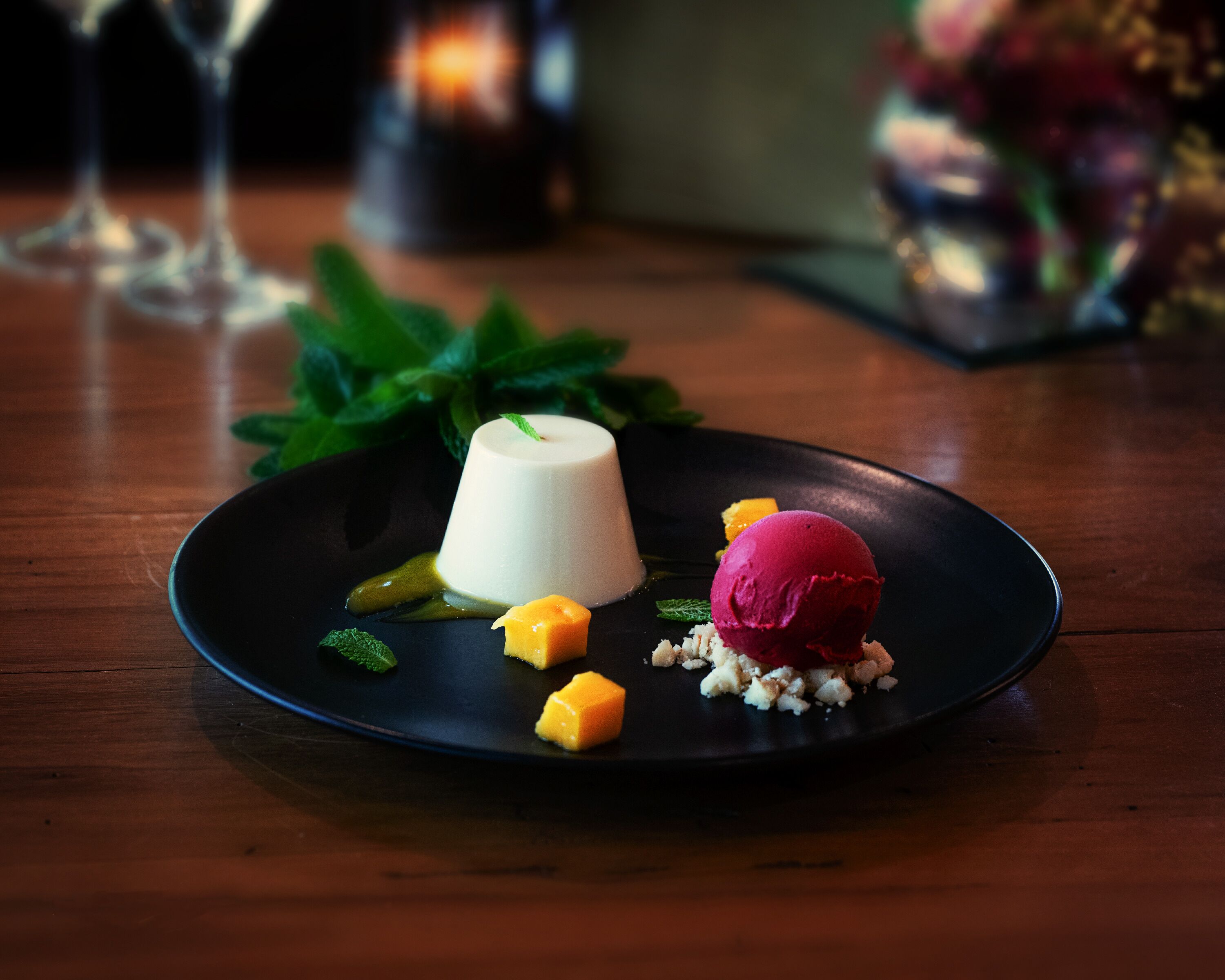 Ein Dessertteller mit Panna Cotta, Mangowürfeln und einer Kugel pinkfarbenem Eis auf dunklem Untertisch.