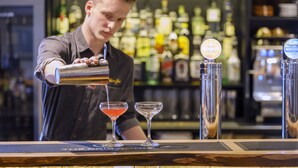 Um bartender servindo uma bebida em uma taça de martini em um bar, com uma variedade de garrafas ao fundo.