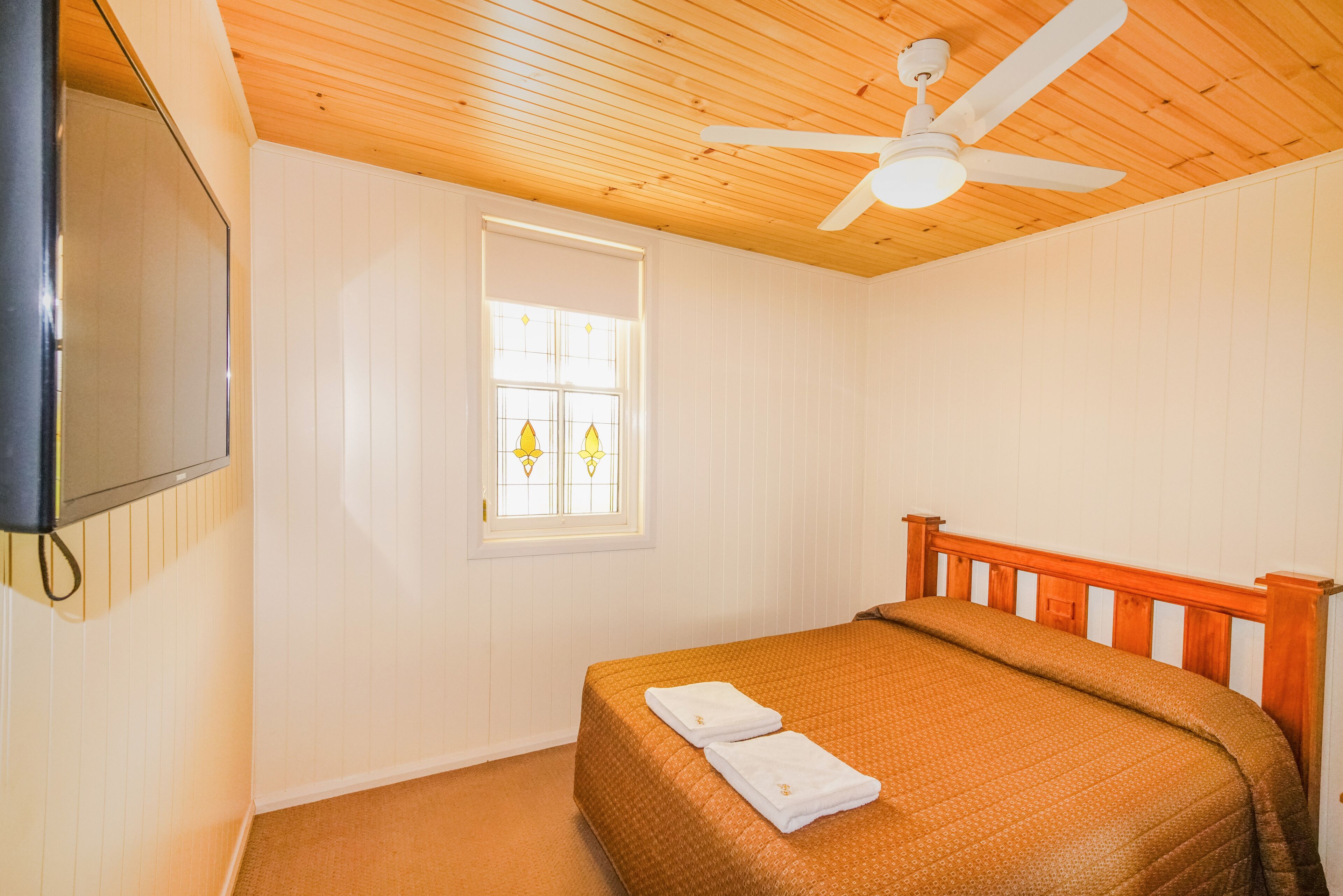 A bedroom with a wooden ceiling, a bed with a brown bedspread, a window with stained glass, and a ceiling fan.