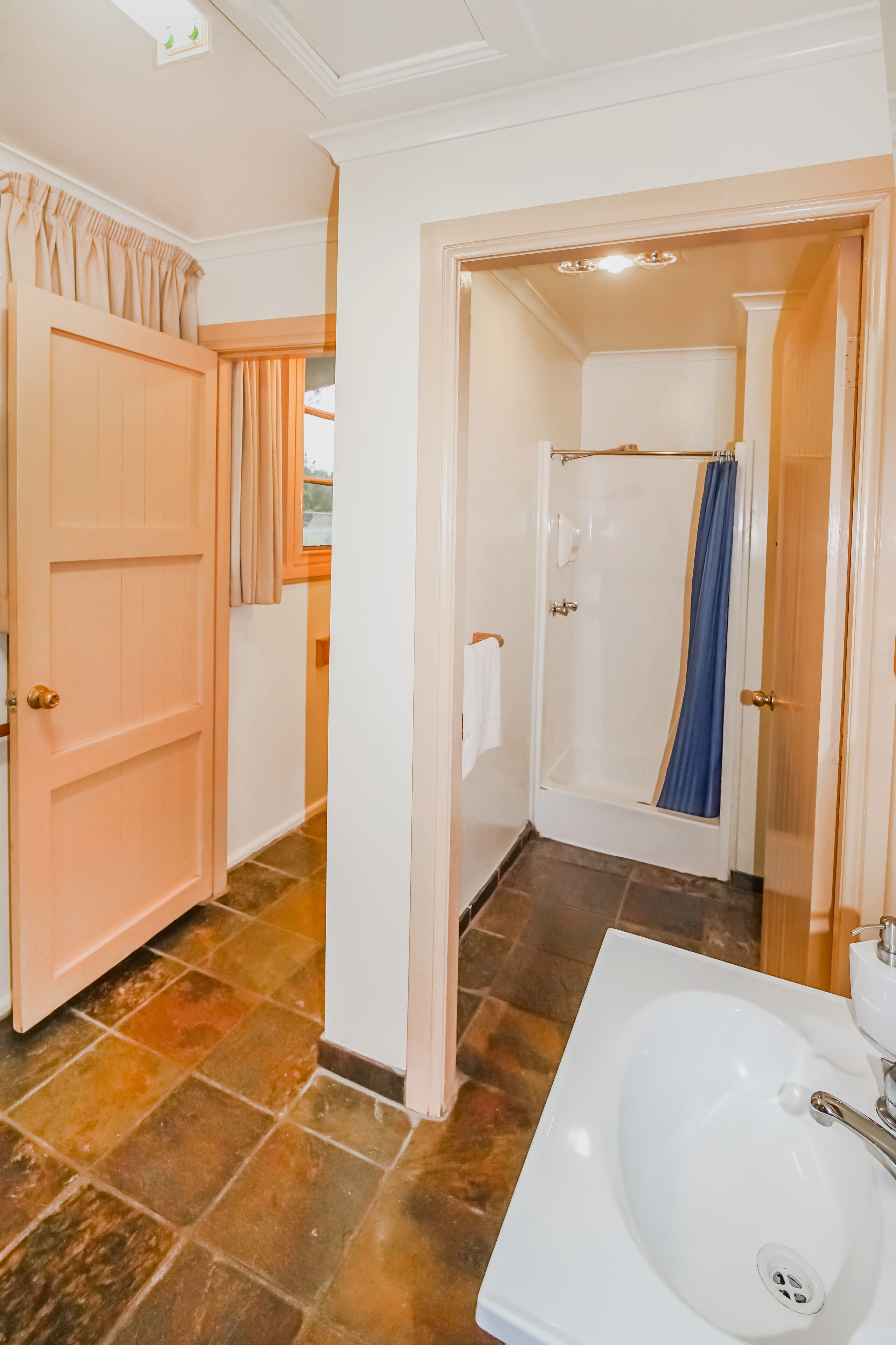 Un bany amb un lavabo blanc, una dutxa amb cortina blava i una porta que condueix a una altra habitació.