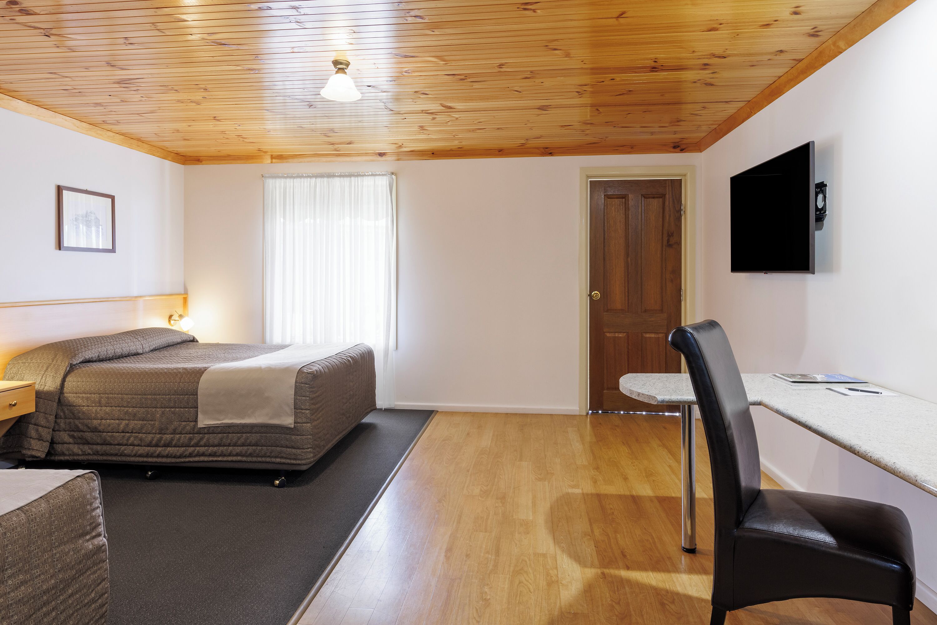 Un dormitorio con una cama, un escritorio, una silla, un televisor, una puerta y una ventana con cortinas.