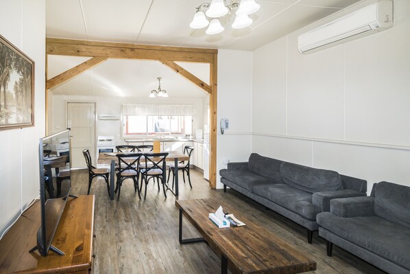 A spacious living area with a wooden floor, a gray sofa, a wooden coffee table, and a dining area with chairs and a table. - Black Gold Motel (Wallerawang)