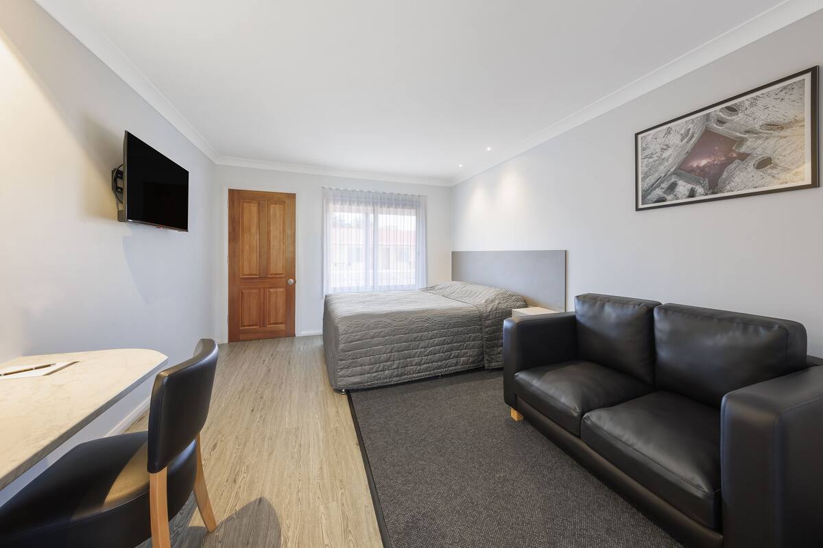 A compact bedroom with a bed, a black leather sofa, a desk with a chair, a TV mounted on the wall, and a window with sheer curtains.