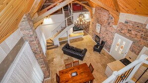 Um amplo quarto estilo loft com teto alto, vigas de madeira aparentes, lareira, sala de jantar com mesa e cadeiras de madeira e escada que leva a um nível superior.