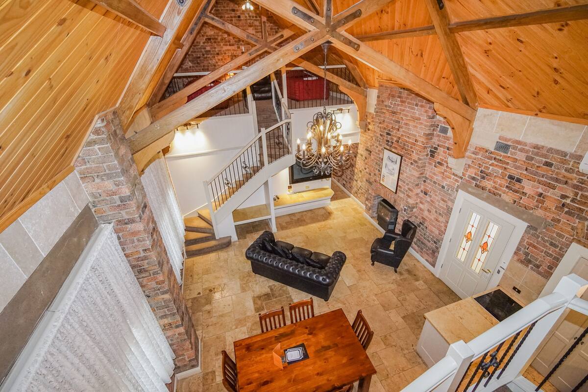 A spacious loft-style room with a high ceiling, exposed wooden beams, a fireplace, a dining area with a wooden table and chairs, and a staircase leading to an upper level.