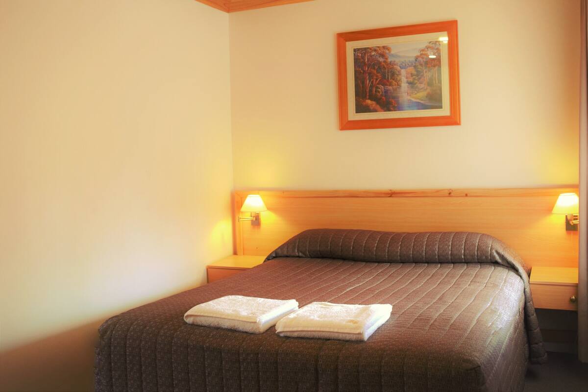 A hotel room with a wooden ceiling, a bed with a brown bedspread, two bedside lamps, and a framed painting on the wall.