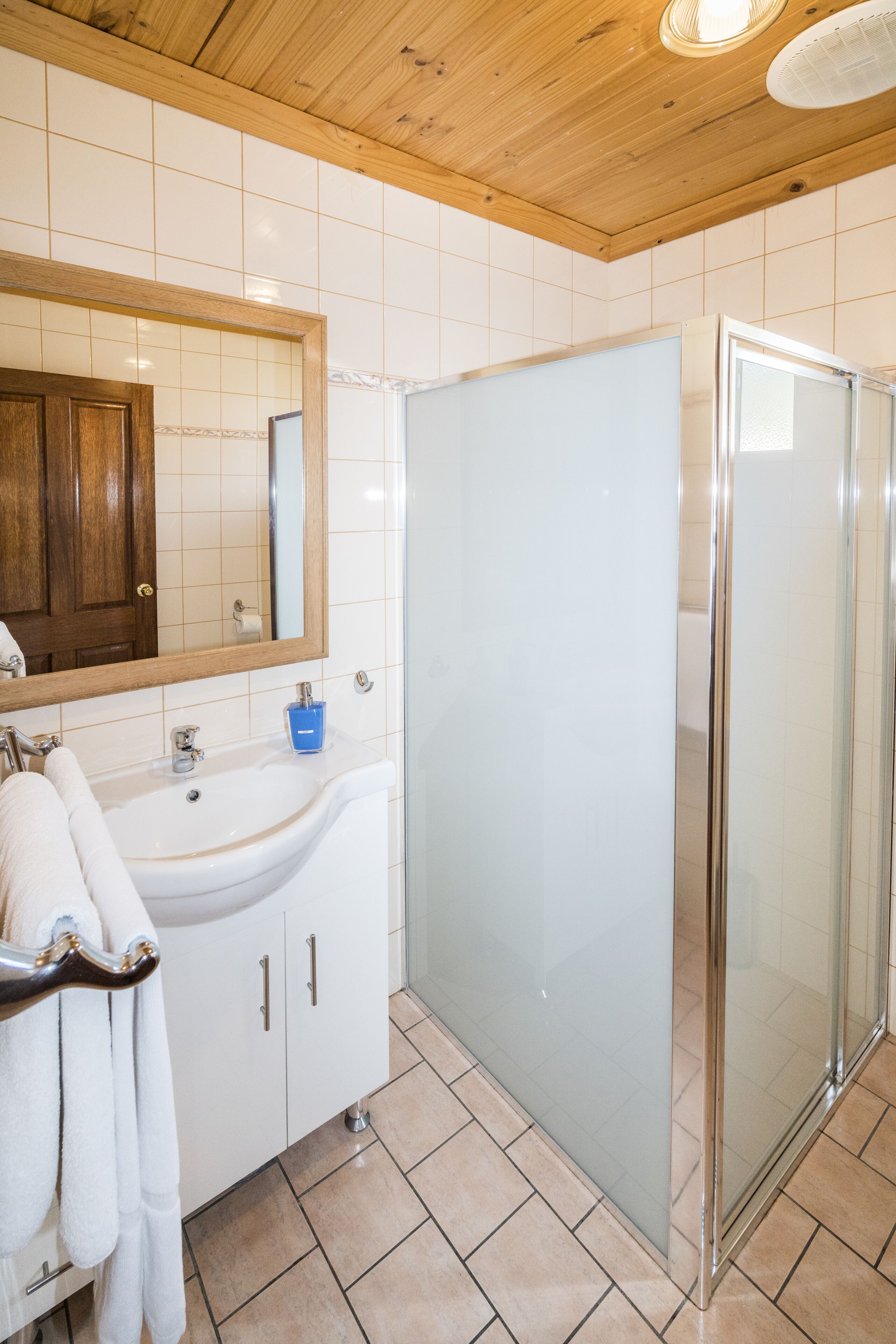 A bathroom with a white sink, a mirror, a glass shower enclosure, and a wooden ceiling.