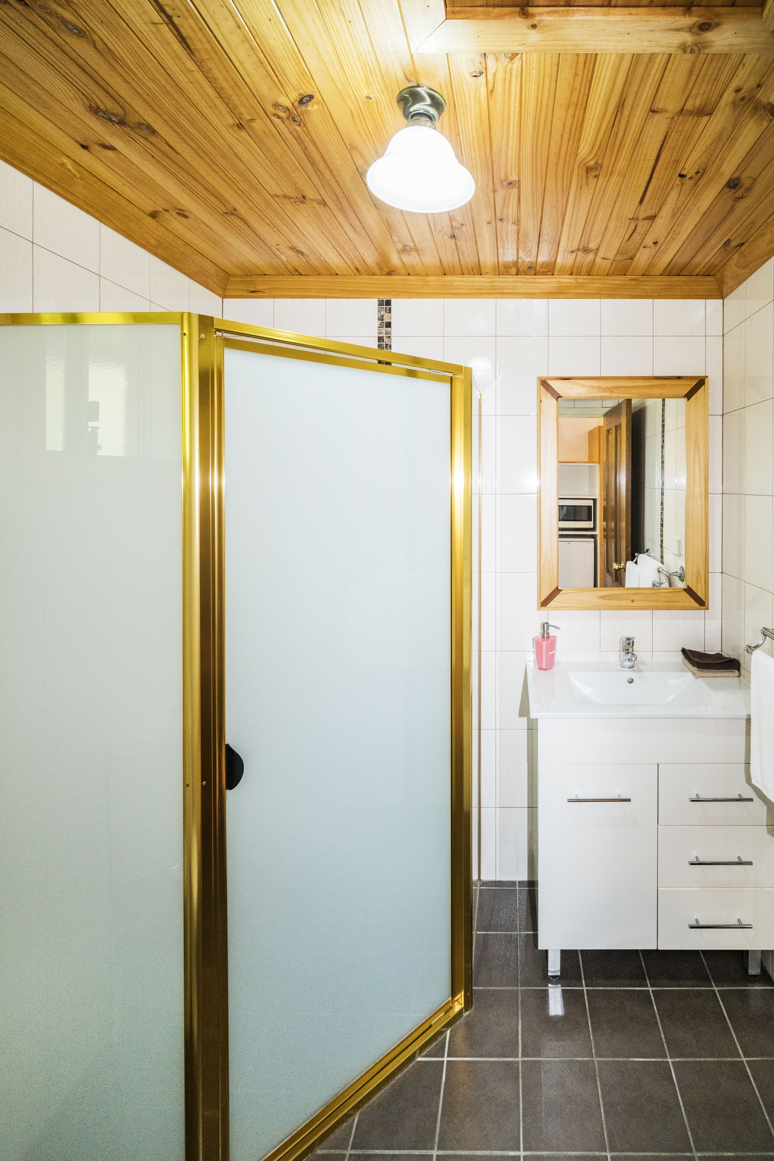 Un baño con techo de madera, baldosas blancas, puerta de vidrio con marco dorado, un lavabo y un espejo.