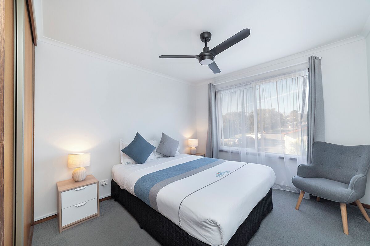 Hotellihuone, jossa on sänky, yöpöytä, lamppu, tuoli ja kattotuuletin.