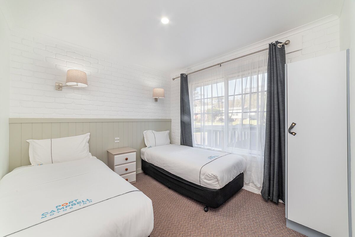 A hotel room with two beds, a white brick wall, a window with curtains, and a door.