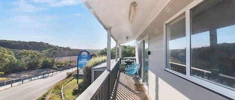 Un balcon donnant sur une route et sur un espace verdoyant, un panneau indiquant un camping près du port, et un ciel dégagé.