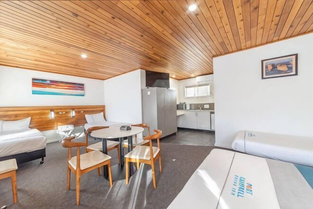 Ein modernes Zimmer mit Holzbalkendecke, einem runden Esstisch mit Stühlen, einem Bett mit Kopfteil, einer Couch, einem Kühlschrank und einer Küchenzeile.