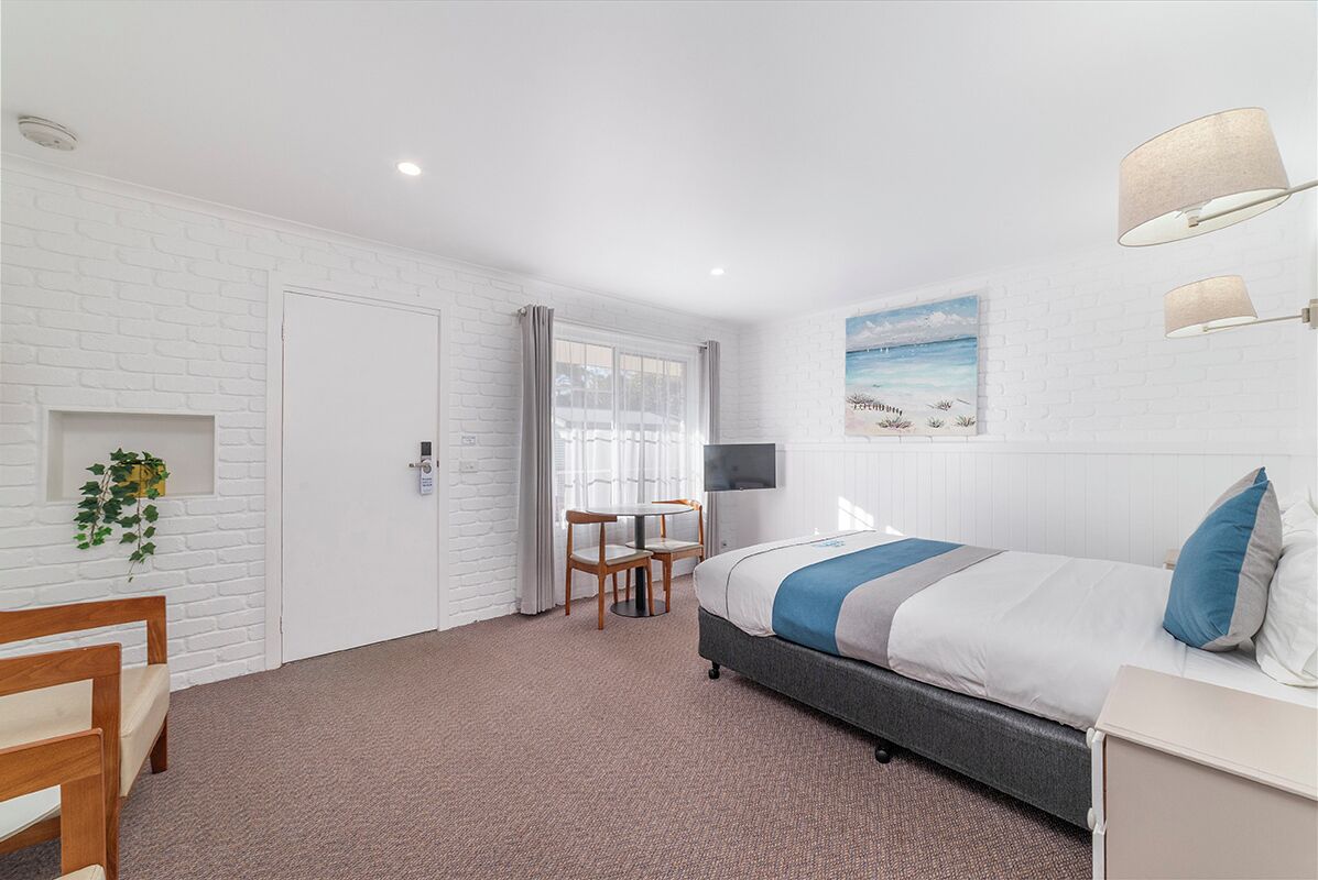 A modern hotel room with a white brick wall, a bed with blue and white bedding, a desk with a chair, a small table with a chair, and a window with curtains.