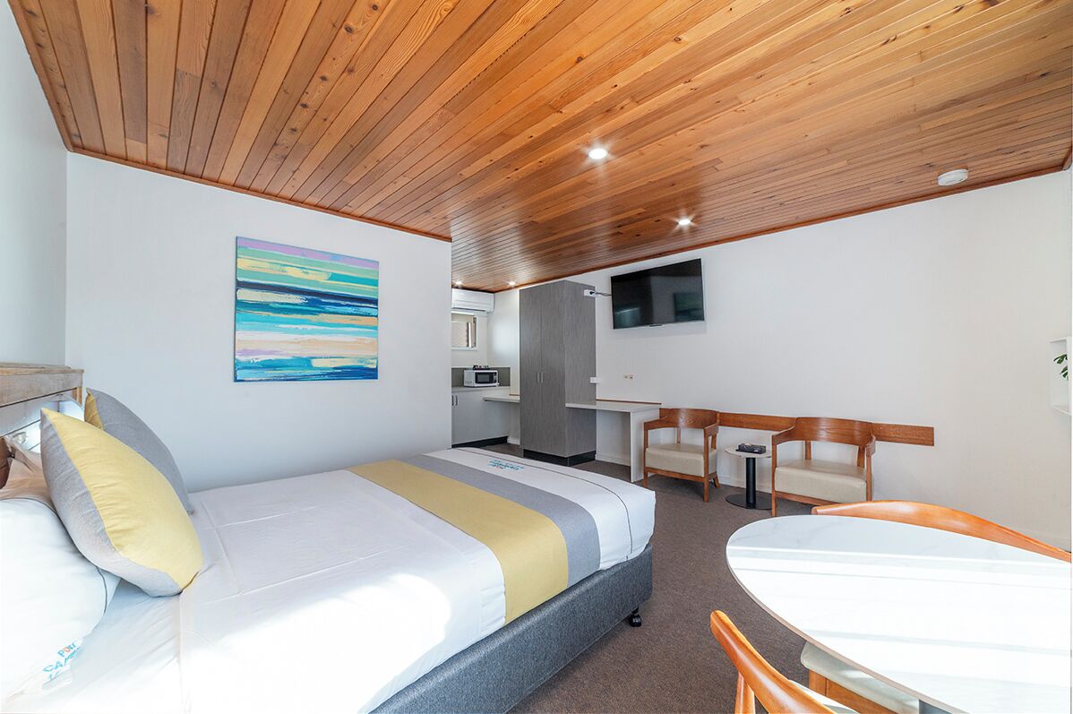 A modern hotel room with a wooden ceiling, a bed with a striped bedspread, a desk with a chair, a television, and a kitchenette.