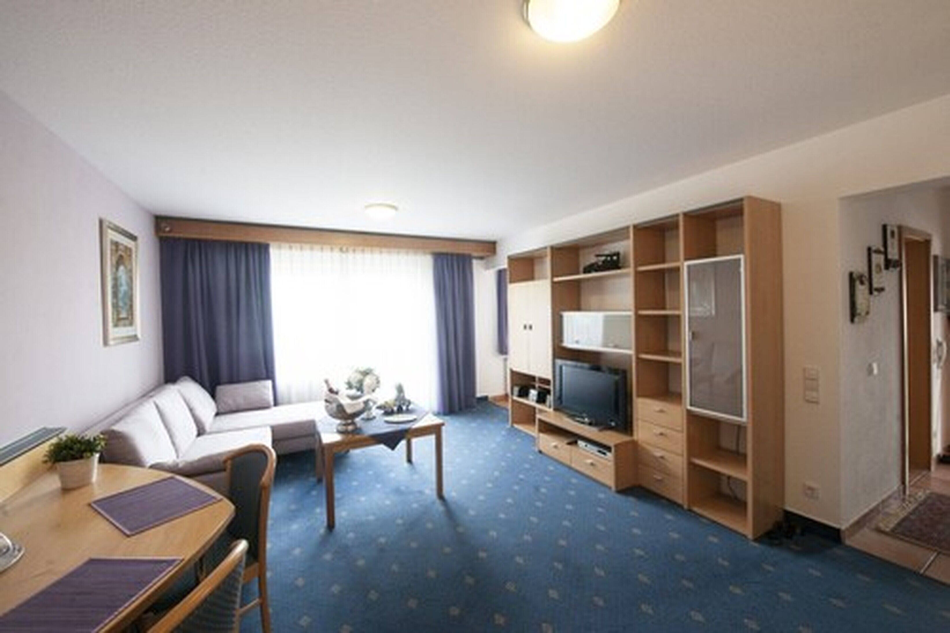 A compact hotel room with a sofa, a dining table, a TV, and built-in storage.