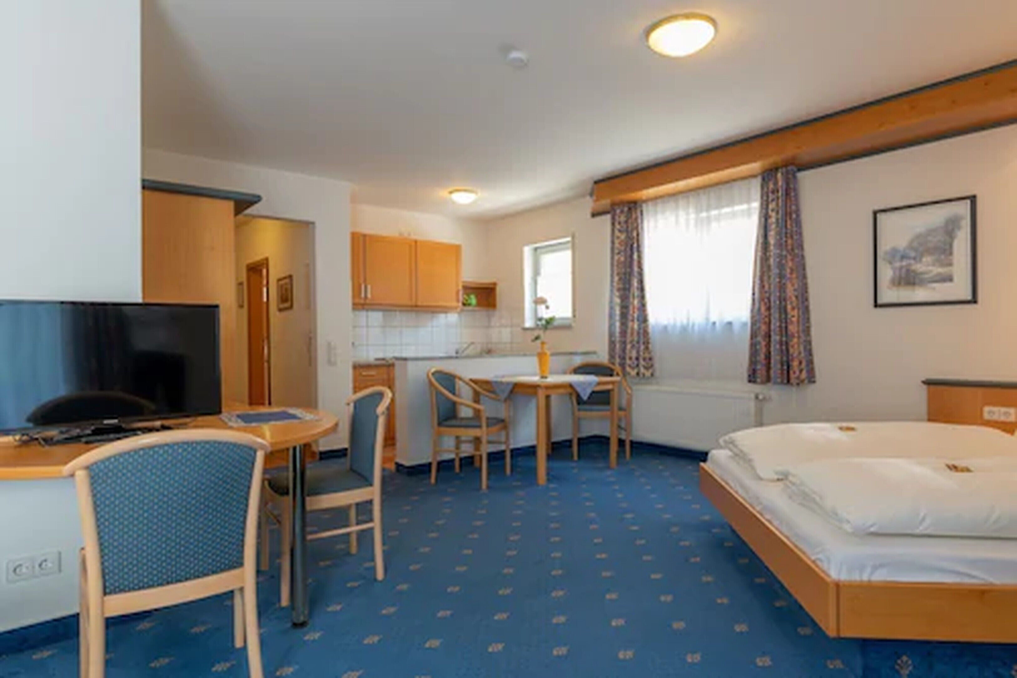 Habitación de hotel con cama, escritorio y silla, televisor, pequeña cocina y ventana con cortinas.