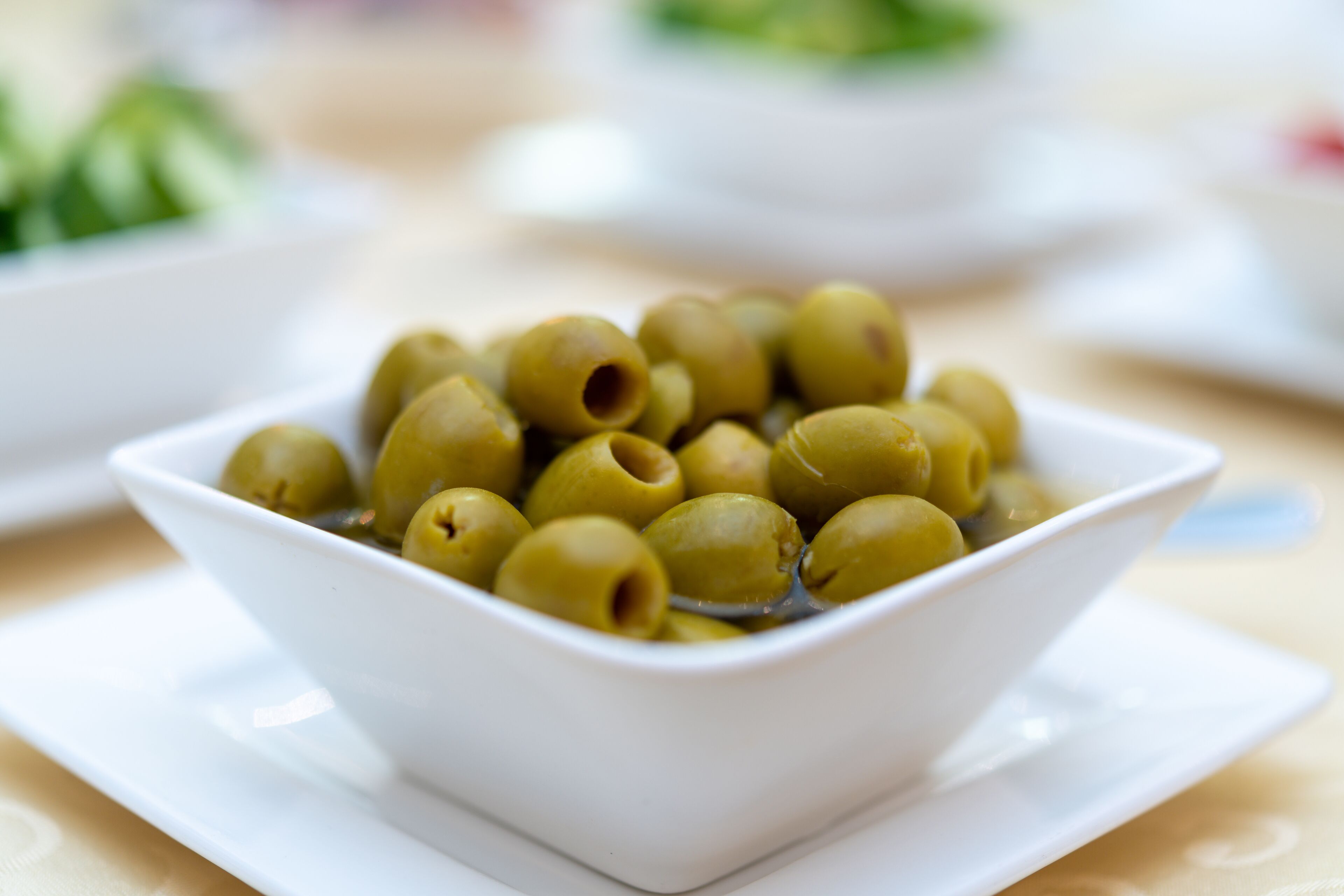 Un bol blanco lleno de aceitunas verdes sobre un plato blanco.