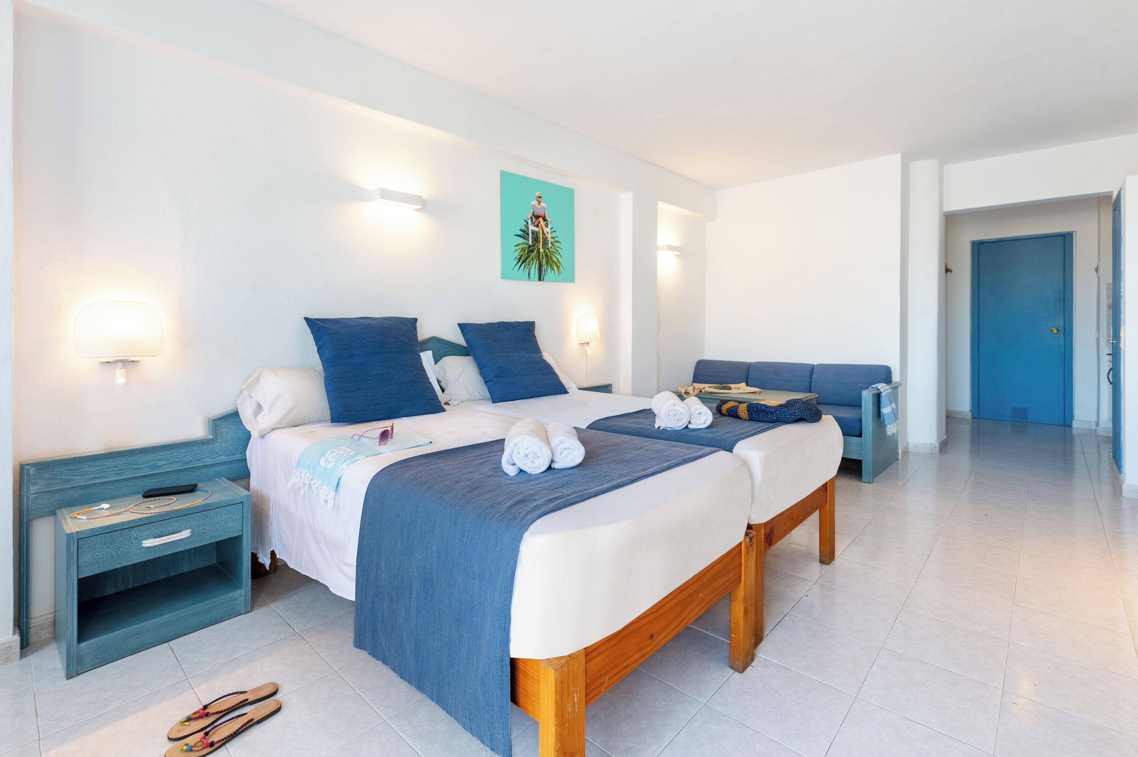 A double bed with blue bedding, a bedside table with a lamp, and a painting on the wall.