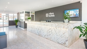 A reception desk with a marble top, a large screen displaying a cityscape, and potted plants. - Aparhotel Vibra Central City - Adults Only (Sant Antoni de Portmany)