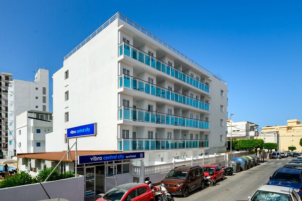 A multi-story building with balconies, a sign for 'Vibra Central City Apart Hotel', and parked cars along the street.
