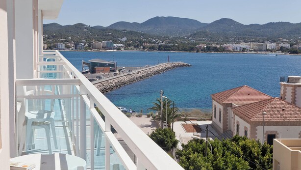 A balcony with a view of a coastal town, a pier, and mountains in the background. - Aparhotel Vibra Central City - Adults Only (Sant Antoni de Portmany)