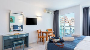 A hotel room with a bed, a desk, a chair, a TV, and a balcony with a view of buildings. - Aparhotel Vibra Central City - Adults Only (Sant Antoni de Portmany)